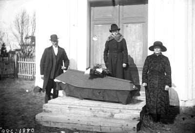 Sörjande föräldrar vid kista med avlidet barn. Oidentifierade. Exteriör: förstugubro.