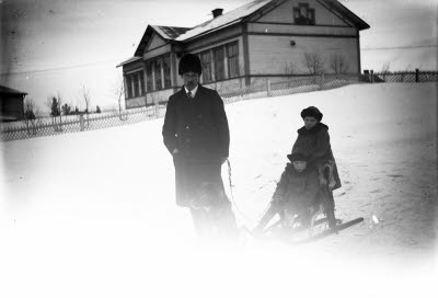 Två barn på sparksläde och en herre med hund. I bakgrunden: Backskolan; Grönskolan, Gällivare.
