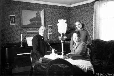 En herre och två damer i hemmiljö. Gammalt piano. Högreståndsmiljö.