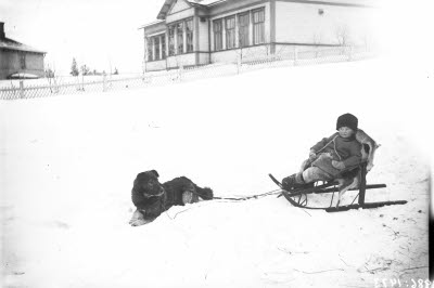 Ett barn på sparksläde och en hund. I bakgrunden: Backskolan; Grönskolan, Gällivare.