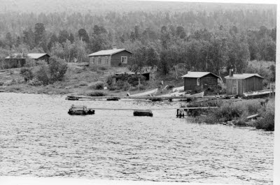 Bebyggelse; stugor vid älvstrand. Vattendrag. Bryggor, båtar.