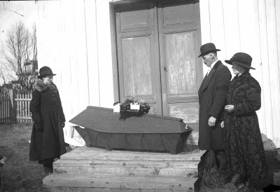 Sörjande föräldrar vid kista med avlidet barn. Oidentifierade. Exteriör: förstugubro.