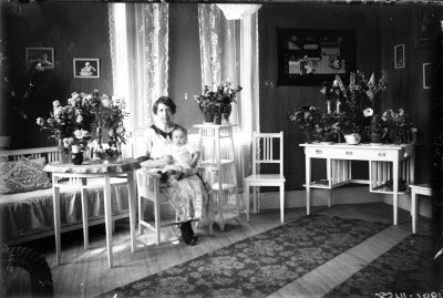 En kvinna och ett barn. Hemmiljö; vardagsrum, piedestal, pinnstol, blomstersmyckade bord och skrivbord.