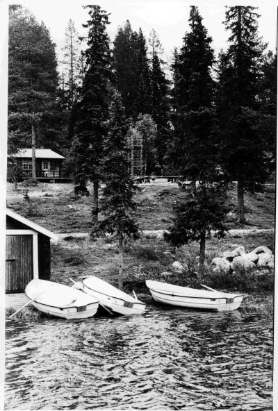 Stug/fritidsområde. Stuga, vatten, båtar, båthus.