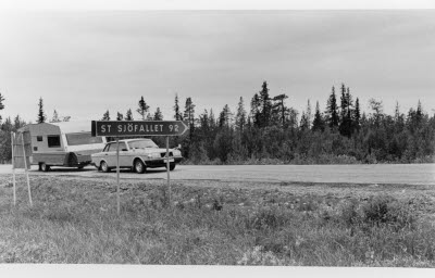 Porjusvägen korsningen Vägen Västerut mot Stora Sjöfallet. Ortnamnsmärke; vägskylt. Personbil med husvagn.