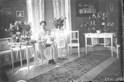 En kvinna och ett barn. Hemmiljö; vardagsrum, piedestal, pinnstol, blomstersmyckade bord och skrivbord.