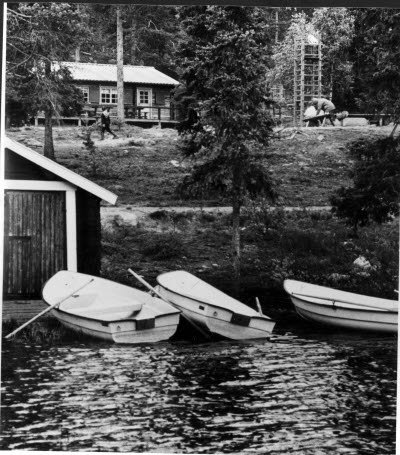 Stug/fritidsområde. Stuga, vatten, båtar, båthus.