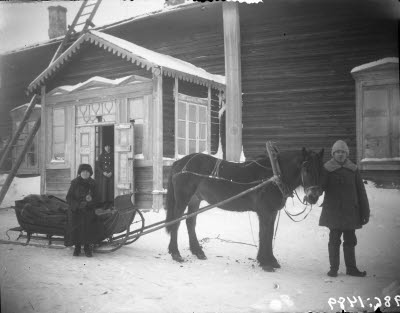 Två damer och en herre. Häst och släde. Förstukvist, gårdsplan.
