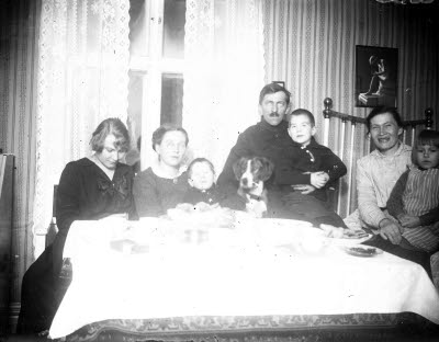 Hemmiljö; interiör, vardagsrum. Tre damer, en herre, tre barn och en hund. Oidentifierade.