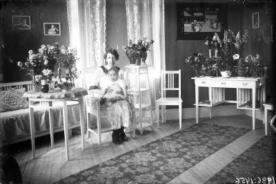 En kvinna och ett barn. Hemmiljö; vardagsrum, piedestal, pinnstol, blomstersmyckade bord och skrivbord.