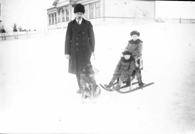 Två barn på sparksläde och en herre med hund. I bakgrunden: Backskolan; Grönskolan, Gällivare.